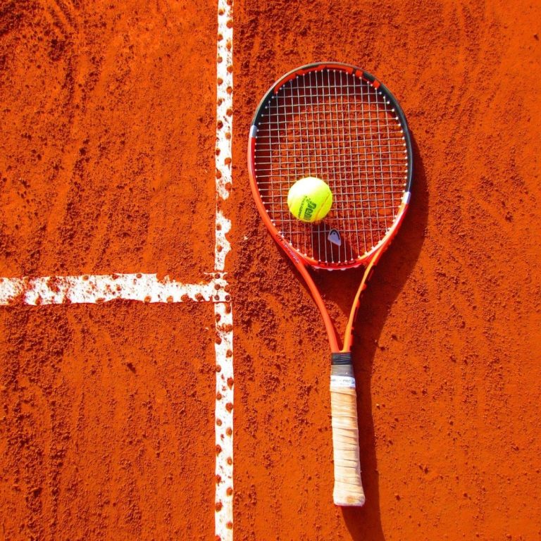 Una raqueta de tenis acostada en cancha de polvo de ladrillo con una pelota arriba.